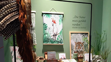 Behind a feathered headdress, illustrations of a cockatoo and of a tiger hang next to the Humboldt quote "Nature must be felt".